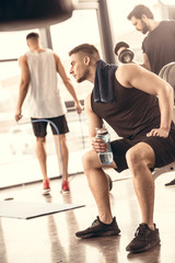 Fototapeta na wymiar side view of handsome sportsmen resting and holding bottle of water in gym