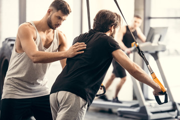 trainer helping sportsman exercising with suspension straps in gym