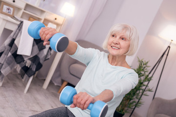 Enjoying workout. Pleasant senior woman grinning while sitting on the floor of the living room and holding a pair of blue dumbbells