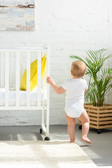 back view of little baby standing at baby crib at home