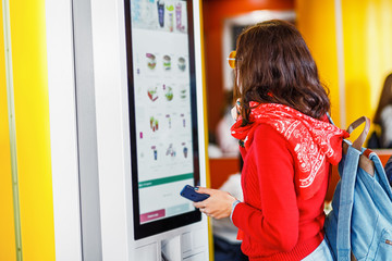 Hipster asian woman orders food at the touch screen self service machine by the electronic menu in the fastfood restaurant