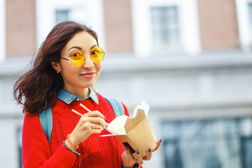 Happy Girl eating asian wok fast food from takeaway paper box at the city street, student or worker...