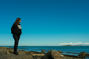 beautiful woman with dark hair dressed black standing on rock formations on coast and looking on ocean. 