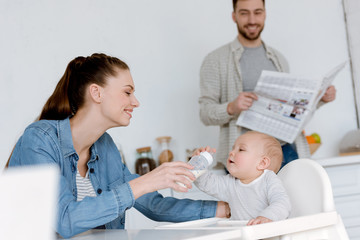 happy mother feeding son with milk in baby bottle on kitchen, father with newspaper behind