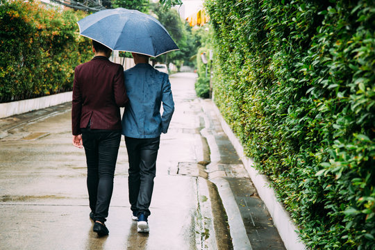 Gay Couple Holding Umbrella And Hands Together. Back Of Homosexual Men Walking In The Rain