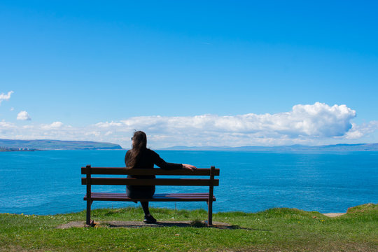 back view of young woman with dark hair sitting on bench against ocean and relaxing. beautiful landscape in portrush northern ireland 