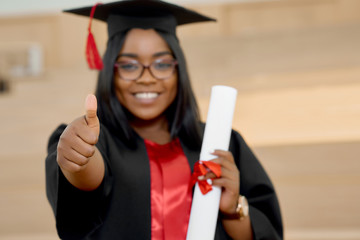 Positive girl graduating from university. Student wearing black and red education gown and keeping diploma.Standing in classroom with wooden cascade desks. Smiling, feeling happy. Blurred focus.