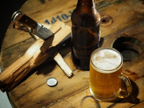 Beer in a glass and a bottle on a wooden table for work, with a knife and a log on the side.