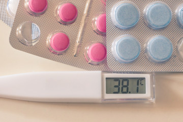 Picture of digital thermometer indicating high temperature and two blisters of blue and pink pills