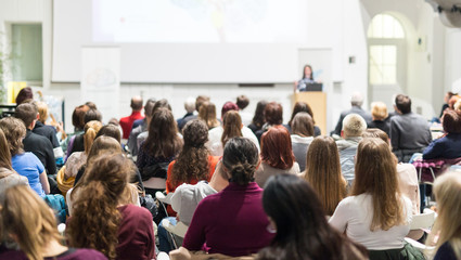 Female speaker giving presentation in lecture hall at university workshop. Audience in conference hall. Rear view of unrecognized participant in audience. Scientific conference event. - obrazy, fototapety, plakaty