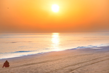 Sunset on the beach with man sitting on sand, long coastline, sun and dramatic sky 