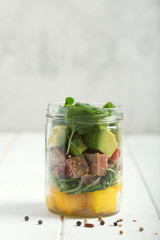 Hawaiian Poke salad with tuna, avocado, mango and vegetables in a jar on a white wooden rustic background
