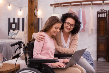 Technology for everyone. Enthusiastic woman embracing handicapped girl who typing