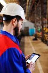 male industrial technician working inside a factory. Man working in a construction helmet with a tablet on the background of a plant