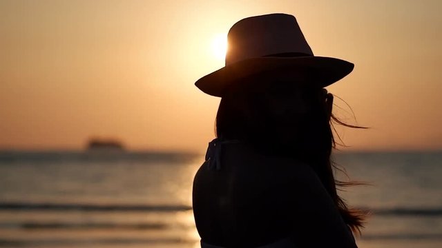 Silhouette of happy asian woman in hat wear white dress standing and looking sunset sea and turned back and looking to camera and smiled. Female traveler enjoy vacation in summer outdoor.