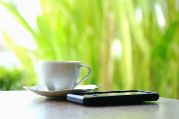 Black coffee in white glass placed behind a black mobile phone on the table near the glass wall in the morning coffee shop.
