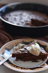 A piece of chocolate cake with prunes lies on a plate decorated with a spoon of sour cream