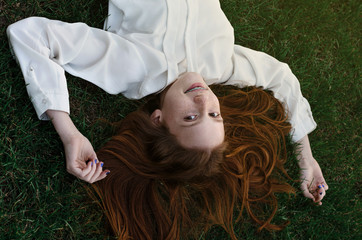 Fototapeta na wymiar Red-haired teen girl in white blouse lies on green grass, arms outstretched and smiling.