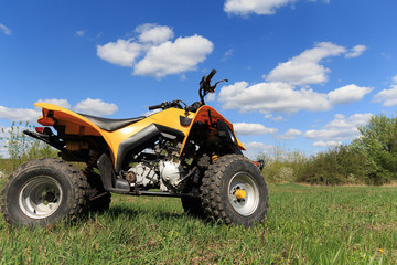 A four-wheeled yellow ATV quad-bike standing idle on the green grass, with trees and a blue sky with clouds on the background © Inna Levchenko