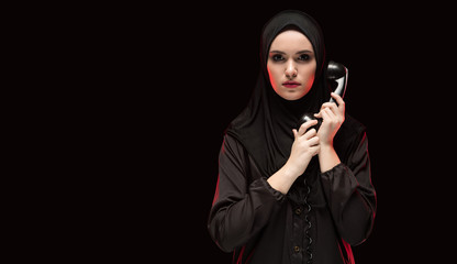 Portrait of beautiful serious scared young woman wearing black hijab holding telephone to call as...