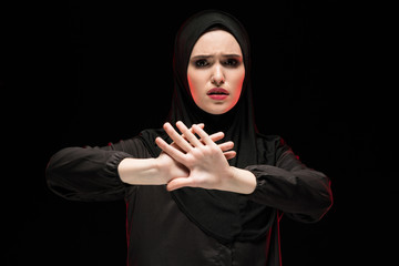 Portrait of young woman in hijab showing stop sign against black background