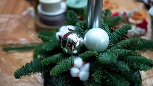 Christmas arrangement: pine tree, pine cones, taper candles, christmas tree toys and cotton flower balls in wicker basket on plastic wrap, close-up. Supply for handcrafted decoration on wooden table.