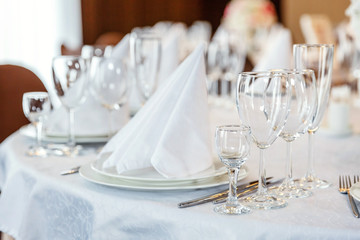 Glasses, flowers, fork, knife, napkin folded in a pyramid, served for dinner in restaurant with cozy interior. Wedding decorations and items for food, arranged by the catering service on a large table