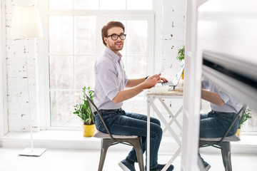 Good working day. Happy bearded man sitting at the table and working on his laptop