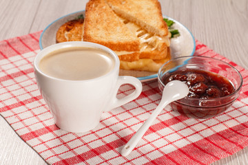 Toasted slices of bread with cheese and green parsley on white plate, cup of coffee, spoon and bowl with strawberry jam on napkin