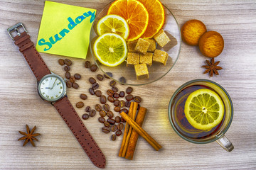 Flat lay. Men's watches with leather strap. Sticker with inscription Sunday. Cup of tea with lemon. Slices of lemon and orange on a saucer. Coffee beans, cookies and pieces of sugar on the table