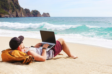 Back view, hipster young man lying on sandy beach leaning on backpack, with laptop computer,...