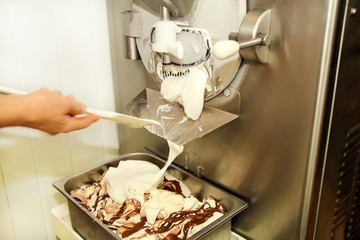Female worker is working on ice cream maker machine. Producing vanilla ice cream with chocolate dressing and it falls into steel container. Industrial preparation of creamy ice cream. Dessert, sweets.