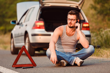 A young man sits on the asphalt, near his broken car and call technical support. Damage on the road