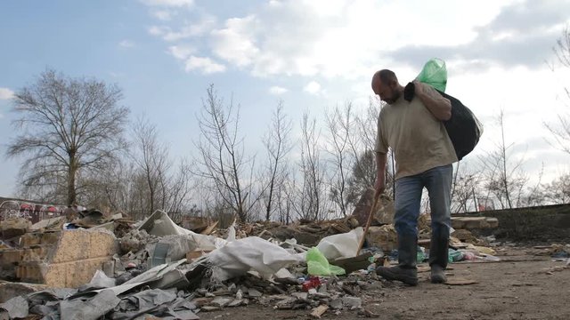 Front view of senior homeless male holding trash bin bags, walking and searching for materials to recycle at garbage dum site. Homelessness and pollution concept. Steadicam shot
