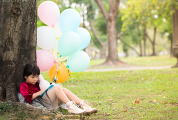 Little girl child read blue book under the big tree. She hugged a white rabbit doll, She has a variety of colored balloons on her side.