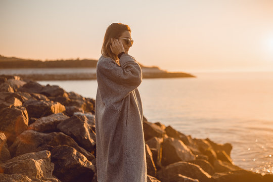 Portrait of woman wearing sunglasses and winter coat standing on rocks looking away at sea, Odessa, Odeska Oblast, Ukraine, Eastern Europe