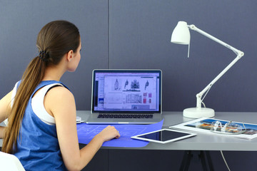 Young woman sitting at the desk with instruments, plan and laptop