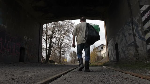 Back view full length of mature unemployed homeless man walking under bridge tunnel in city. Poor male collecting plastic to earn money with recycling. Steadicam stabilized shot.