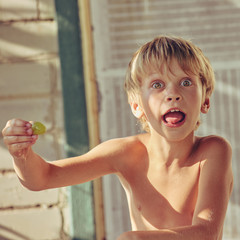 Happy little boy having fun and grimacing while eating grape, image with square aspect ratio and...