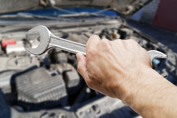 Closeup mans hand holds a wrench against blurred background of the engine in the open hood of the car