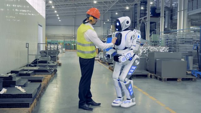 Male factory worker is remotely controlling the robot and they are walking together