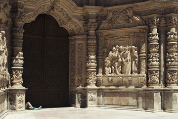 Picture of the facade of a huge church with some statues of women talking and whispering each other and two manly legs in green shorts and sneakers.
