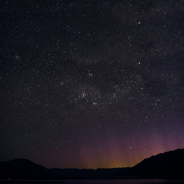 Night picture of a minimal landscape of the stars and Southern Cross in New Zealand. The purple, yellow, red and orange lights emerging behind the mountains came from Queenstown.