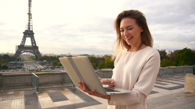 Young female person chatting by laptop near Eiffel Tower in slow motion. Concept of modern technology and tourism in France. Pretty woman wears blouse and has blonde hair.