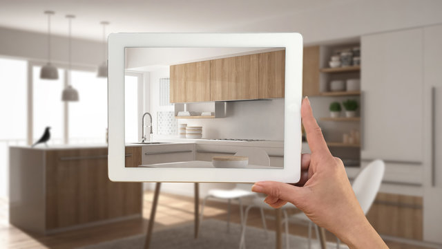 Hand holding tablet showing real finished minimalist white and wooden kitchen. Modern blurred kitchen in the background, architecture interior design presentation