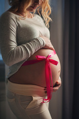 A pregnant woman touches her stomach. On his stomach a pink bow is tied. A woman expects that a girl will be born