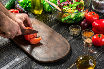 Man's hand cuts ripe red tomatoes for summer healthy vegetable salad on a wooden board, vertical