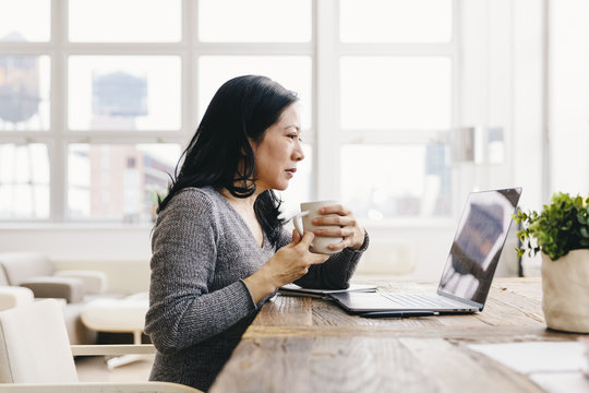 Businesswoman holding coffee cup while using laptop in office