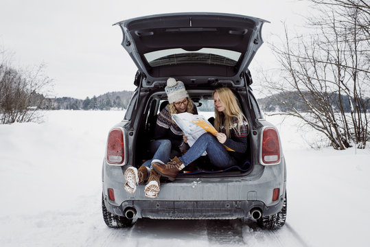 Female friends reading map while sitting in car trunk during winter