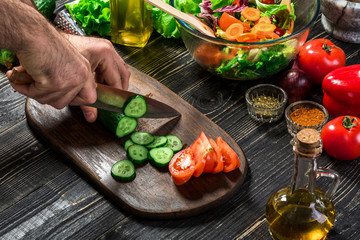 The man is cutting the cucumber in the kitchen in his house to prepare salad along with eating dinner.